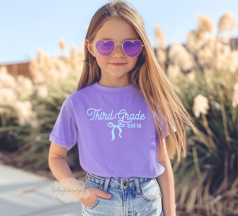 May include: A young person wearing heart-shaped sunglasses and a lavender t-shirt. The shirt has the words "Third Grade SOFIA" printed on it in a light blue script font with a bow graphic. The person is wearing blue jeans.