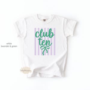 May include: A white short-sleeved t-shirt featuring a graphic print of vertical lavender stripes. Centered on the stripes is the text club ten in a green cursive font, positioned above a green line-drawn bow graphic on the front of the shirt.