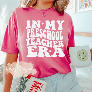 May include: A pink t-shirt with the text "IN MY PRESCHOOL TEACHER ERA" in white retro-style lettering. The shirt is worn by a person with blonde hair and a necklace. The person is holding a white mug with a smiley face on it.