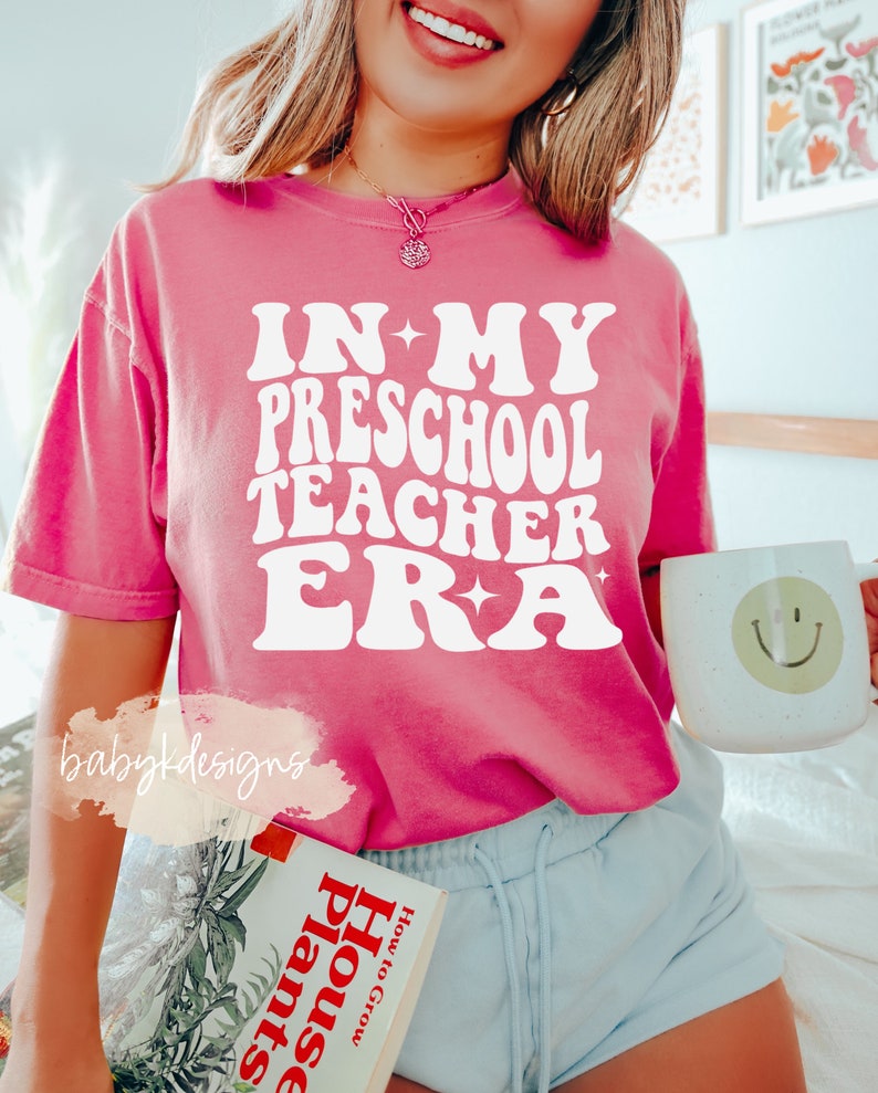 May include: A pink t-shirt with the text "IN MY PRESCHOOL TEACHER ERA" in white retro-style lettering. The shirt is worn by a person with blonde hair and a necklace. The person is holding a white mug with a smiley face on it.