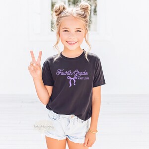 May include: A young girl wearing a black t-shirt with the words "Fourth Grade" and the name "Kaitlyn" in a purple script. The girl is wearing denim shorts and making a peace sign with her fingers.