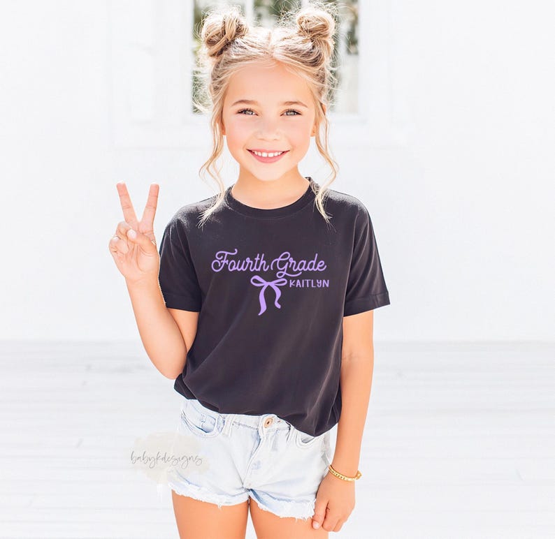 May include: A young girl wearing a black t-shirt with the words "Fourth Grade" and the name "Kaitlyn" in a purple script. The girl is wearing denim shorts and making a peace sign with her fingers.