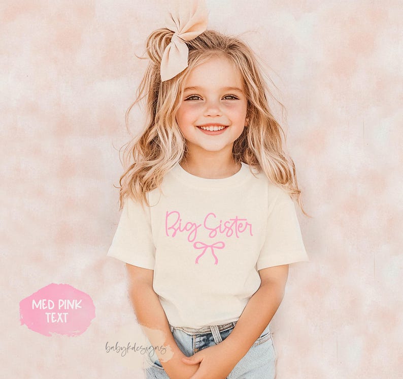 May include: A young girl wearing a cream-colored t-shirt with the words "Big Sister" in pink script and a pink bow graphic. The girl is smiling and wearing a large bow in her hair. The text "MED PINK TEXT" is also visible.