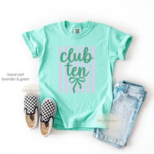 May include: A mint green short-sleeve t-shirt featuring vertical lavender stripes and the text "club ten" in dark green script, accompanied by a dark green bow graphic. The shirt is displayed with a pair of light blue denim shorts and checkered slip-on shoes.