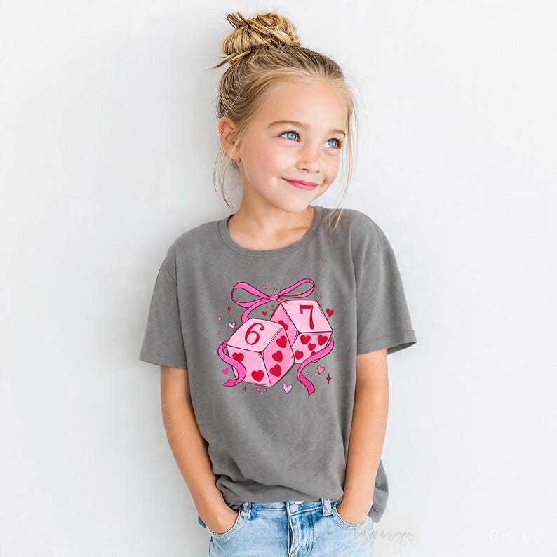 May include: A gray t-shirt featuring a graphic of two pink dice with the numbers 6 and 7, decorated with hearts and a pink ribbon. The shirt is worn by a young girl with blonde hair.