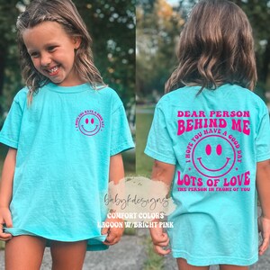 May include: A turquoise blue t-shirt with a pink smiley face on the front and the text "Dear person behind me I hope you have a good day! Lots of love the person in front of you" on the back.