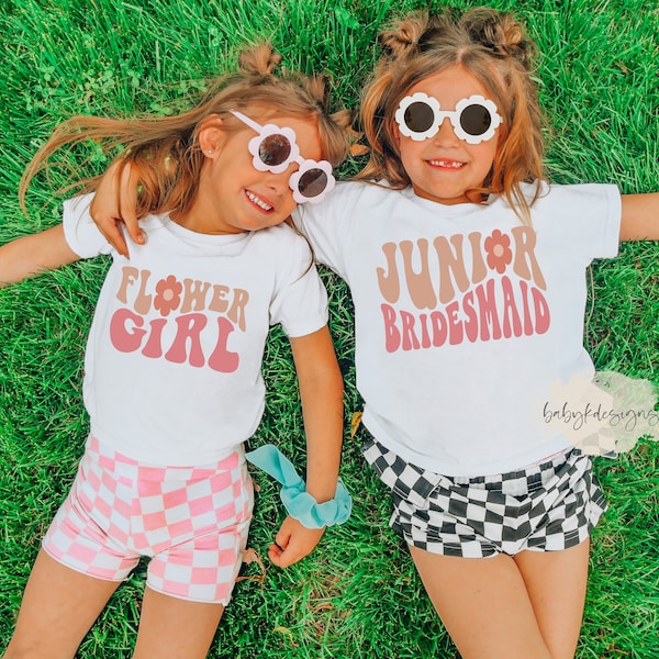 May include: Two white short-sleeve t-shirts featuring retro-style text. One shirt displays the words Flower Girl in pink and tan with a flower icon, paired with pink and white checkered shorts. The other shirt reads Junior Bridesmaid in similar colors, paired with black and white checkered shorts.