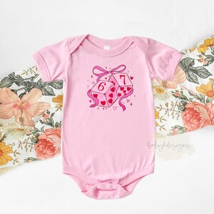May include: A light pink baby onesie with a graphic of two dice, one showing a 6 and the other a 7. The dice are decorated with hearts and tied with a pink ribbon. The onesie is short-sleeved.