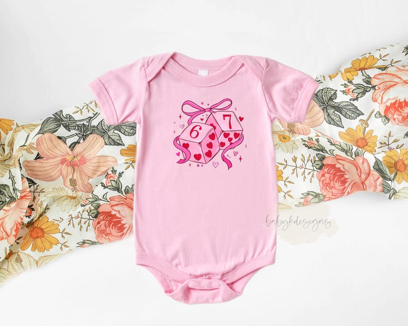 May include: A light pink baby onesie with a graphic of two dice, one showing a 6 and the other a 7. The dice are decorated with hearts and tied with a pink ribbon. The onesie is short-sleeved.