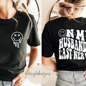 May include: Black t-shirt with a white dripping smiley face on the front and the text "On my husband's last nerve" in a retro font on the back.