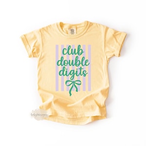 May include: A pale yellow short-sleeve t-shirt featuring a graphic design with vertical light purple stripes. The text "club double digits" is printed in green cursive script, positioned above a green bow illustration on the front of the shirt.