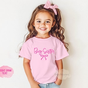 May include: A young girl wearing a light pink t-shirt with the words "Big Sister" in bright pink script. A pink bow is printed below the text. The girl is smiling and wearing a matching pink bow in her hair.