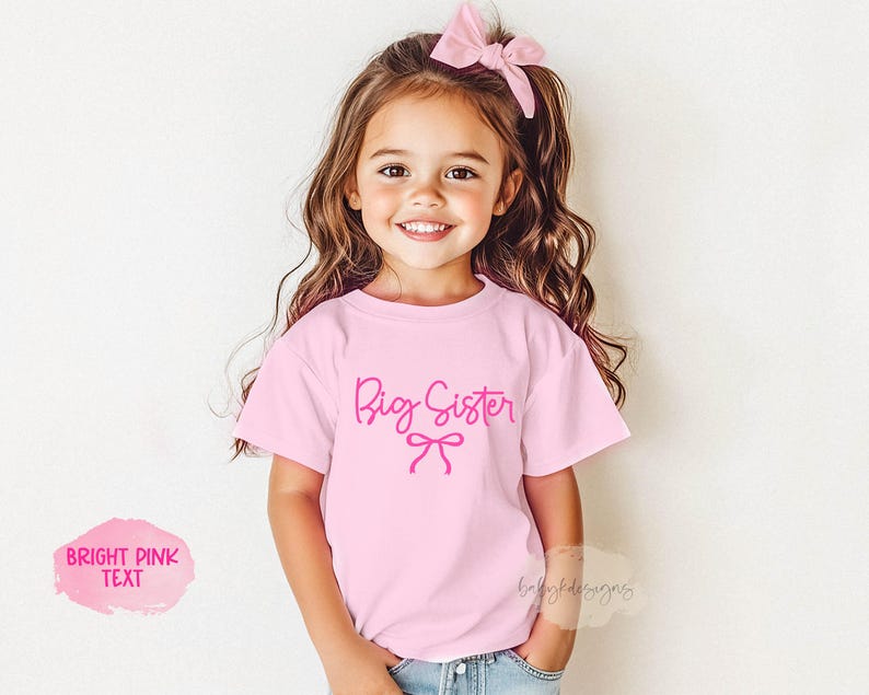 May include: A young girl wearing a light pink t-shirt with the words "Big Sister" in bright pink script. A pink bow is printed below the text. The girl is smiling and wearing a matching pink bow in her hair.