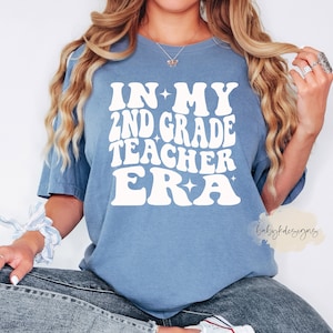 May include: A blue t-shirt with white text that reads "In My 2nd Grade Teacher Era" with stars on either side of the text. The text is in a retro font.