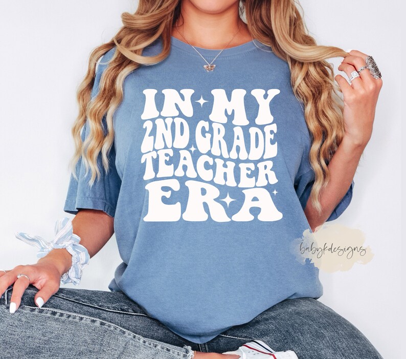 May include: A blue t-shirt with white text that reads "In My 2nd Grade Teacher Era" with stars on either side of the text. The text is in a retro font.