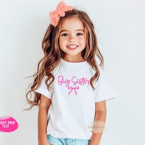 May include: A white t-shirt with the words "Big Sister" in bright pink script and a bow graphic. The shirt is worn by a young girl with long brown hair and a pink bow. The text "Bright Pink Text" is in a pink oval.
