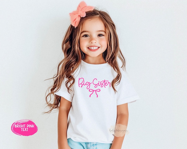 May include: A white t-shirt with the words "Big Sister" in bright pink script and a bow graphic. The shirt is worn by a young girl with long brown hair and a pink bow. The text "Bright Pink Text" is in a pink oval.