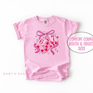 May include: A light pink t-shirt featuring a graphic of two dice with the numbers 6 and 7, decorated with hearts and a pink ribbon. The text "Comfort Colors Youth & Adults Sizes" is in a white oval. The shirt has rolled-up sleeves.