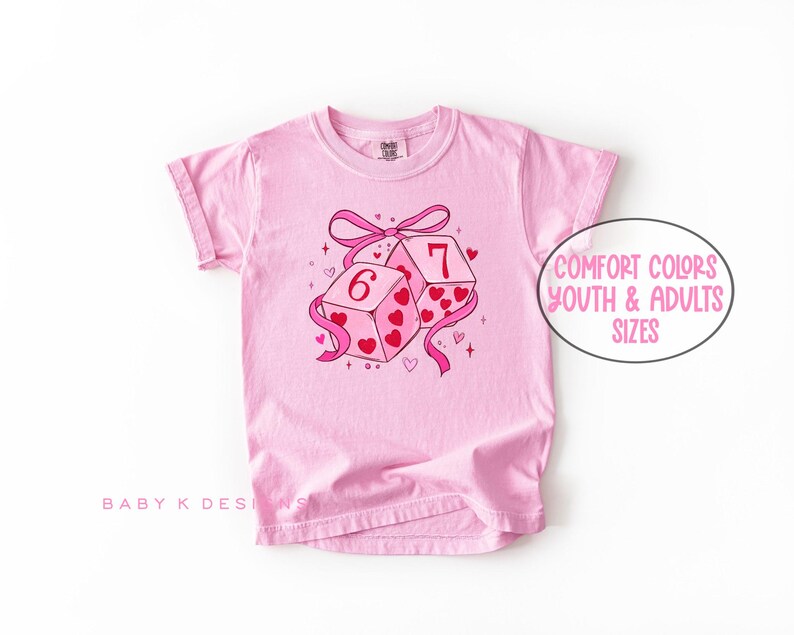 May include: A light pink t-shirt featuring a graphic of two dice with the numbers 6 and 7, decorated with hearts and a pink ribbon. The text "Comfort Colors Youth & Adults Sizes" is in a white oval. The shirt has rolled-up sleeves.