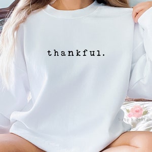 Op de afbeelding: Een witte trui met de tekst "thankful" in zwarte letters op de borst. De tekst is in een eenvoudig lettertype. De trui heeft een klassieke ronde hals en is geschikt voor dagelijks gebruik. De stof lijkt zacht.