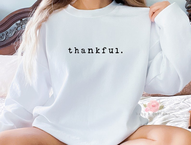Op de afbeelding: Een witte trui met de tekst "thankful" in zwarte letters op de borst. De tekst is in een eenvoudig lettertype. De trui heeft een klassieke ronde hals en is geschikt voor dagelijks gebruik. De stof lijkt zacht.