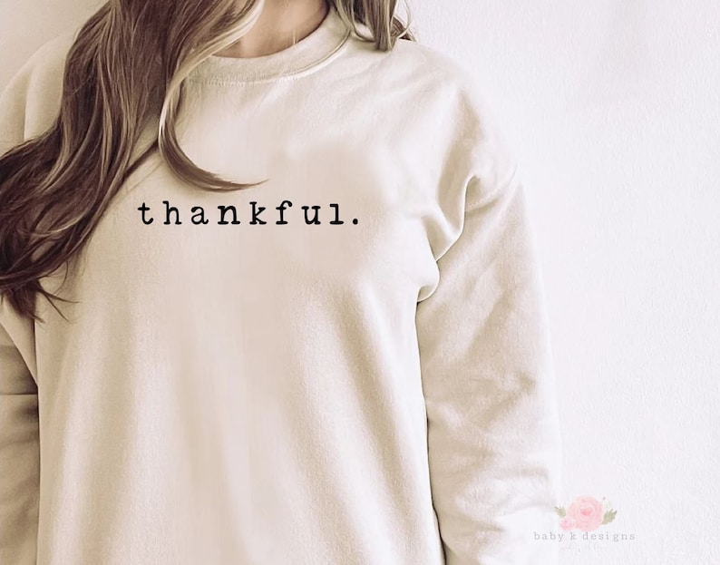 Op de afbeelding: Cr&egrave;mekleurige sweatshirt met het woord "thankful" in zwart gedrukt, gecentreerd bovenaan. De sweatshirt heeft een ronde hals en lange mouwen. De afbeelding is een close-up van het bovenlichaam.