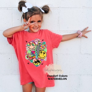 May include: A coral-coloured t-shirt with a colourful graphic of smiley faces, sunglasses, a disco ball, and the words "Hello Summer." The design includes a watermelon slice and a heart with palm trees. The shirt is labelled "Comfort Colors Watermelon."