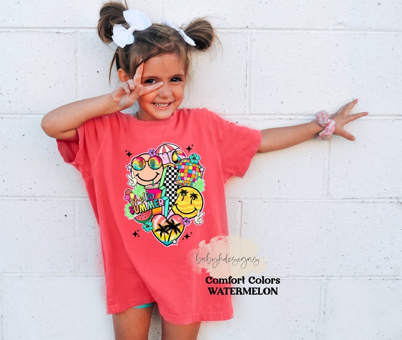 May include: A coral-coloured t-shirt with a colourful graphic of smiley faces, sunglasses, a disco ball, and the words "Hello Summer." The design includes a watermelon slice and a heart with palm trees. The shirt is labelled "Comfort Colors Watermelon."