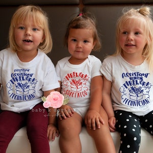 May include: Three young children wearing white tops. Two t-shirts say "Assistant to the Firework Technician" in blue, and the onesie says "Firework Spectator" in red. The children are sitting and smiling.