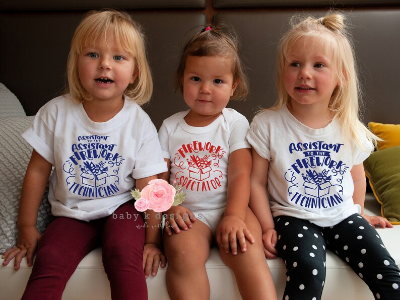 May include: Three young children wearing white tops. Two t-shirts say "Assistant to the Firework Technician" in blue, and the onesie says "Firework Spectator" in red. The children are sitting and smiling.