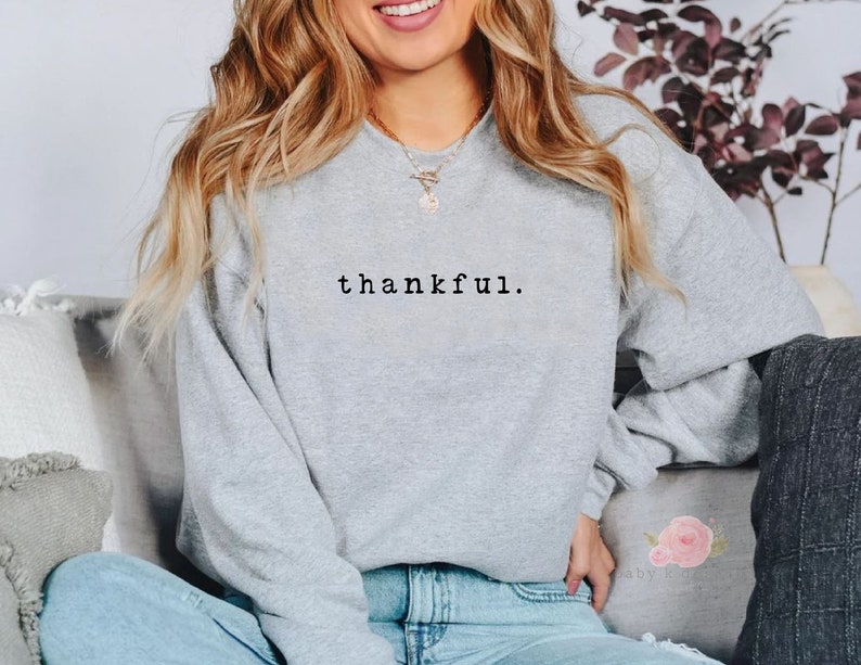 Op de afbeelding: Een lichtgrijs sweatshirt met het woord "thankful" in zwarte letters. De sweatshirt heeft een ronde hals en lange mouwen. De afbeelding toont een persoon die het sweatshirt draagt.