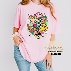 May include: A light pink t-shirt featuring a vibrant graphic. The design incorporates smiley faces, a disco ball, a watermelon slice, and the text "hello summer". The shirt is a comfort colors brand.