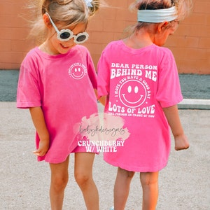 May include: Pink t-shirt with a white graphic that says "Dear Person Behind Me I Hope You Have A Good Day Lots Of Love The Person In Front Of You". The graphic also includes a smiley face.