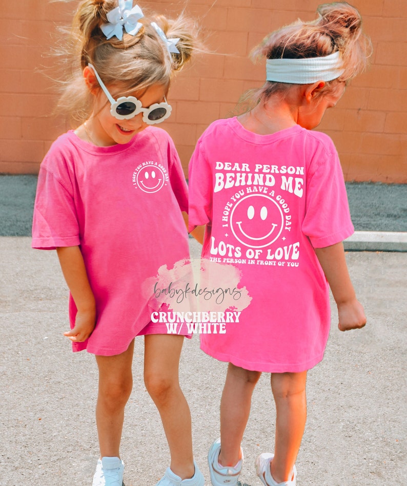 May include: Pink t-shirt with a white graphic that says "Dear Person Behind Me I Hope You Have A Good Day Lots Of Love The Person In Front Of You". The graphic also includes a smiley face.