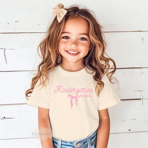 May include: A young girl wearing a cream-colored t-shirt with the word "Kindergarten" and the name "Olivia" printed in pink. A pink bow graphic is below the text. The girl is smiling and wearing a beige bow in her hair.