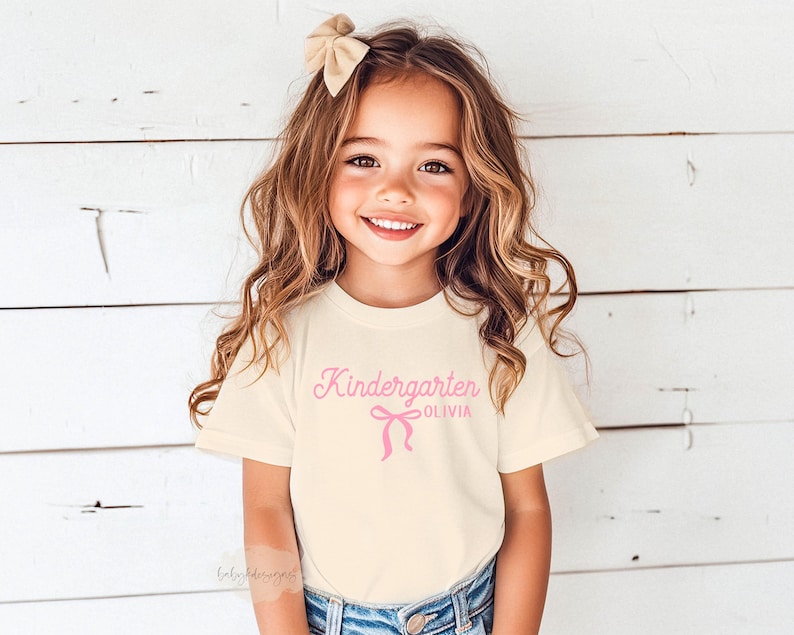 May include: A young girl wearing a cream-colored t-shirt with the word "Kindergarten" and the name "Olivia" printed in pink. A pink bow graphic is below the text. The girl is smiling and wearing a beige bow in her hair.