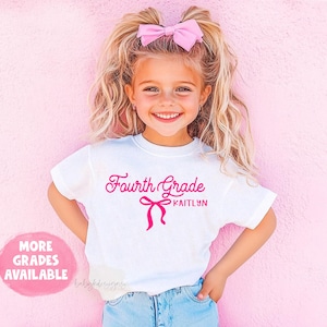May include: White t-shirt with the text "Fourth Grade KAITLYN" in pink script and a bow graphic. The girl is wearing a pink bow in her hair. A pink circle with the text "MORE GRADES AVAILABLE" is in the lower left corner.