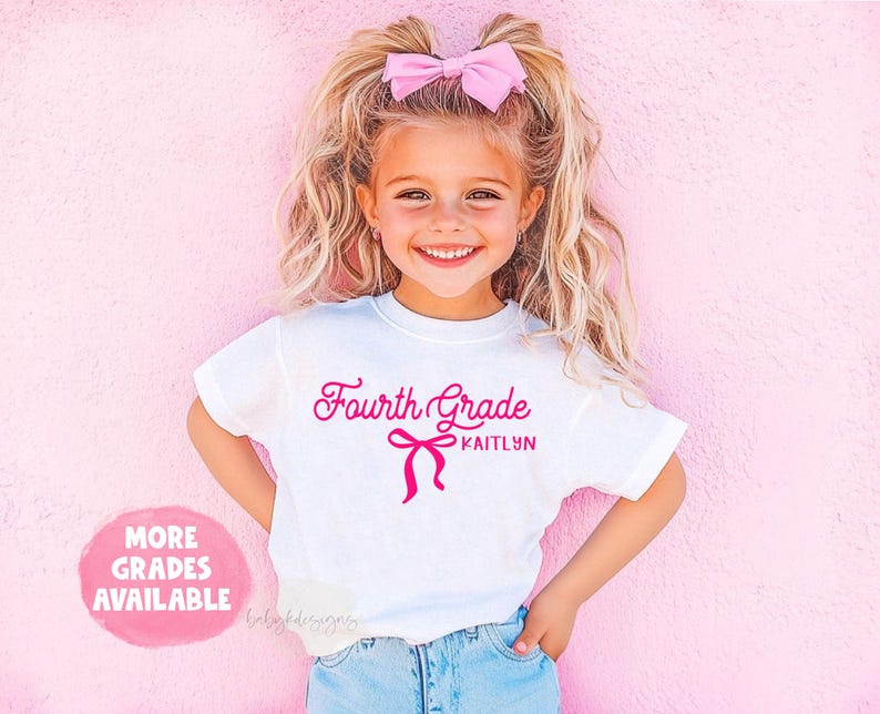 May include: White t-shirt with the text "Fourth Grade KAITLYN" in pink script and a bow graphic. The girl is wearing a pink bow in her hair. A pink circle with the text "MORE GRADES AVAILABLE" is in the lower left corner.