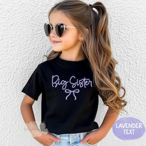 May include: A young girl wearing a black t-shirt with the lavender text "Big Sister" and a bow design. She is also wearing heart-shaped sunglasses. The shirt is paired with blue jeans.