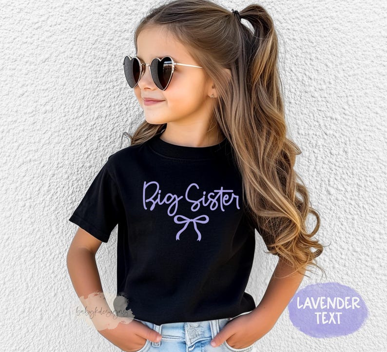 May include: A young girl wearing a black t-shirt with the lavender text "Big Sister" and a bow design. She is also wearing heart-shaped sunglasses. The shirt is paired with blue jeans.