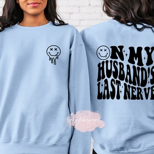 May include: A light blue sweatshirt with a black dripping smiley face on the front and the text "On my husband's last nerve" on the back.
