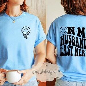 May include: A light blue t-shirt with a black dripping smiley face on the front and the text "On My Husband's Last Nerve" in a retro font on the back.