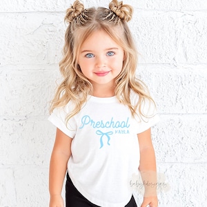 May include: A young child wearing a white t-shirt with the word "Preschool" in light blue script, a bow graphic, and the name "Kayla". The child has blonde hair and is smiling.