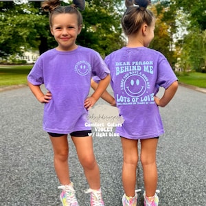May include: Purple short-sleeved t-shirt with a light blue smiley face graphic on the front and the text "Dear person behind me I hope you have a good day Lots of love the person in front of you" on the back.