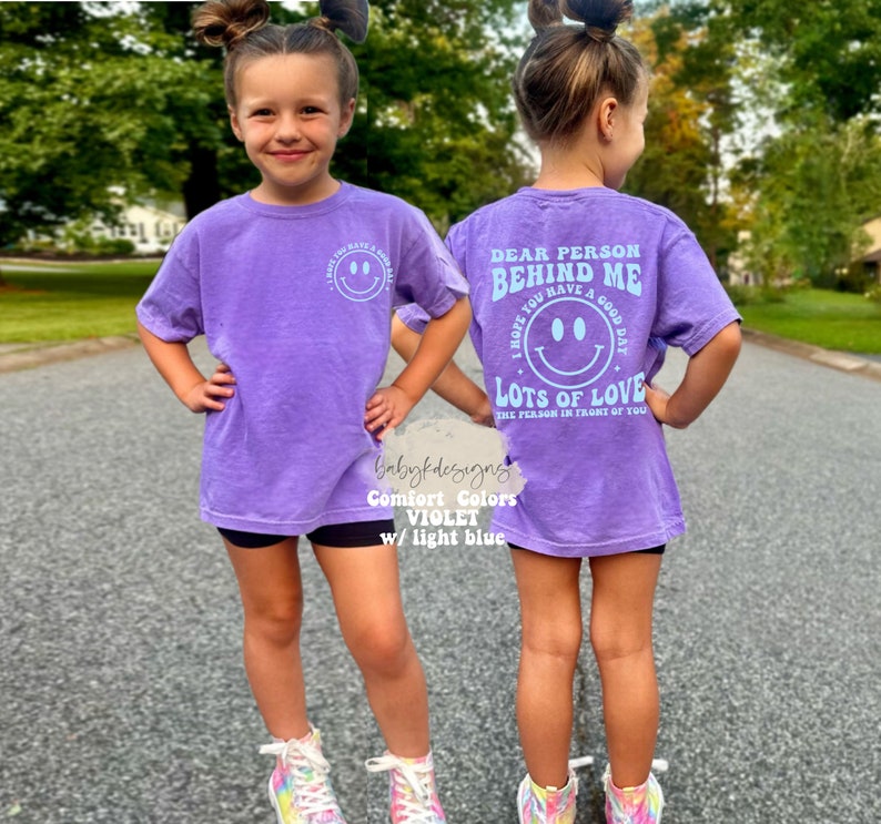 May include: Purple short-sleeved t-shirt with a light blue smiley face graphic on the front and the text "Dear person behind me I hope you have a good day Lots of love the person in front of you" on the back.
