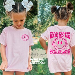 May include: Pink t-shirt with a pink smiley face and the text "I hope you have a good day" on the front and the text "Dear person behind me, I hope you have a good day. Lots of love, the person in front of you" on the back.