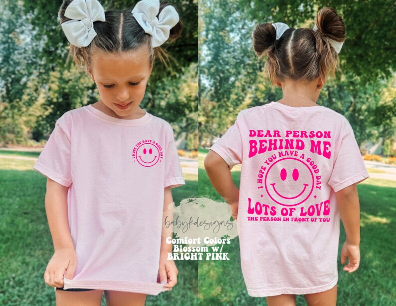 May include: Pink t-shirt with a pink smiley face and the text "I hope you have a good day" on the front and the text "Dear person behind me, I hope you have a good day. Lots of love, the person in front of you" on the back.