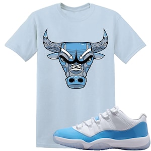 May include: A pale blue t-shirt with a blue and grey bull head design. The image also shows white and light blue trainers with a translucent sole. The shirt and trainers share a similar colour palette.