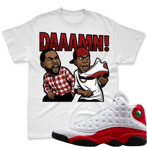 May include: White t-shirt with a cartoon graphic of two men reacting to a red and white sneaker. The word "DAAAMN!" is printed in red above the cartoon. A red and white Air Jordan 13 sneaker is in the foreground.