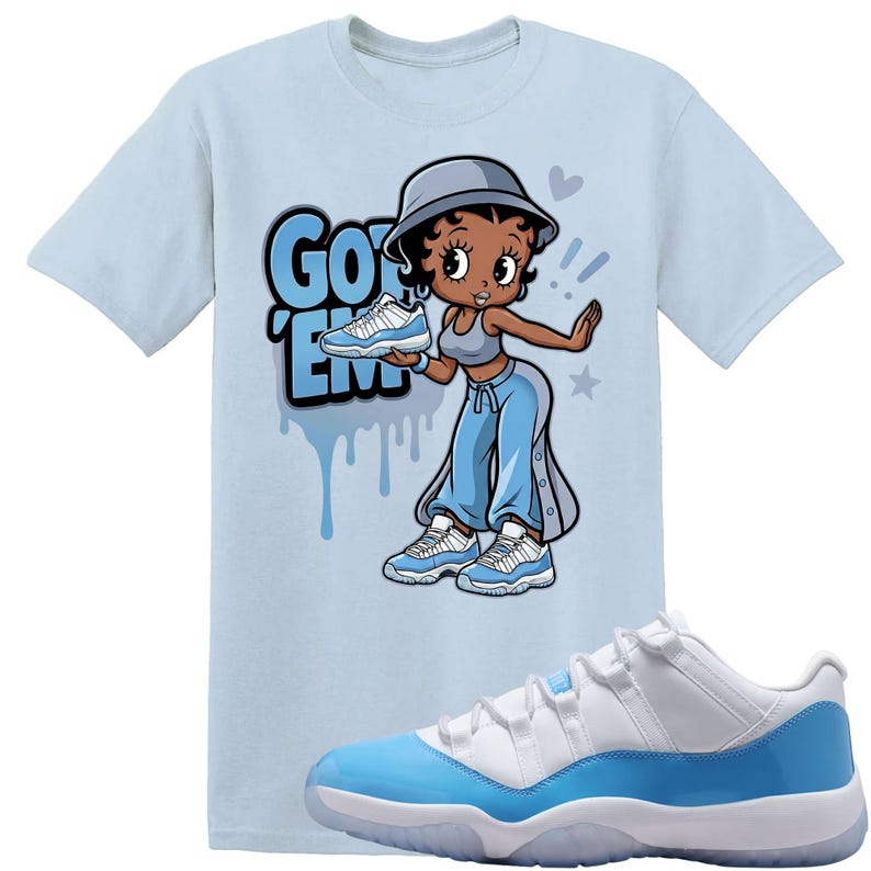 Op de afbeelding: Lichtblauw T-shirt met een cartoonfiguur die een paar blauw-witte sneakers vasthoudt. De tekst "Got 'Em" is in blauw weergegeven. Een paar bijpassende sneakers staat op de voorgrond.